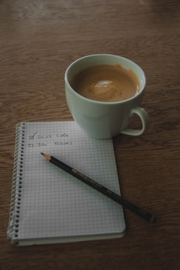 A white cup of coffee sits on a wooden table next to a spiral-bound notebook with a pencil resting on top. The notebook page contains a checklist with one item marked as complete and another yet to be checked.