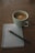 A white cup of coffee sits on a wooden table next to a spiral-bound notebook with a pencil resting on top. The notebook page contains a checklist with one item marked as complete and another yet to be checked.
