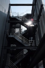 An industrial-style metal staircase with multiple flights and platforms is situated in an open-air setting between two buildings. The sunlight filters dramatically through the stairs, casting shadows and highlighting the geometric lines and railings.
