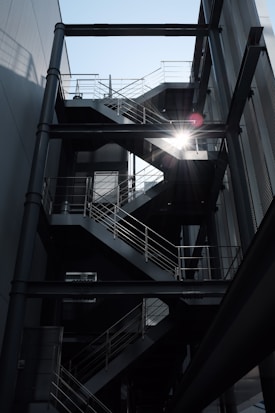 An industrial-style metal staircase with multiple flights and platforms is situated in an open-air setting between two buildings. The sunlight filters dramatically through the stairs, casting shadows and highlighting the geometric lines and railings.