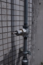 A metal pipe with a valve is mounted on a tiled wall. The wall has a pattern of small, square tiles. The surface appears weathered, suggesting exposure to outdoor elements.