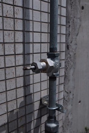 A metal pipe with a valve is mounted on a tiled wall. The wall has a pattern of small, square tiles. The surface appears weathered, suggesting exposure to outdoor elements.