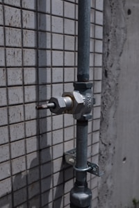 A metal pipe with a valve is mounted on a tiled wall. The wall has a pattern of small, square tiles. The surface appears weathered, suggesting exposure to outdoor elements.
