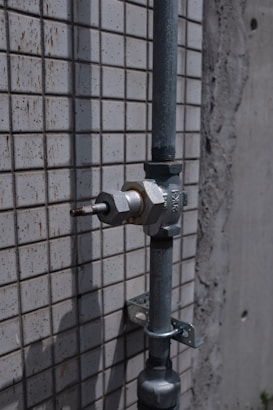 A metal pipe with a valve is mounted on a tiled wall. The wall has a pattern of small, square tiles. The surface appears weathered, suggesting exposure to outdoor elements.