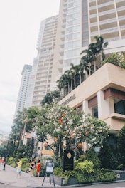 A tall, modern high-rise building with multiple balconies, surrounded by lush green trees and neatly trimmed shrubs. People in casual clothing are walking along the sidewalk in front of the building, and a large sign is prominently displayed at the entrance.