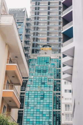 Tall modern buildings with glass facades rise closely together, with one building labeled 'PRIME' and another 'VENUE.' The architecture features a mix of geometric designs, balconies, and varying shades of blue and gray glass. The perspective looks upward, capturing the narrow space between two adjacent structures on the left and right sides.