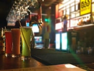 Three tall glasses filled with colorful beverages are arranged on a bar counter. The background features dim ambient lighting, with glowing signs and a row of hanging glasses creating a cozy atmosphere. The focus on the drinks contrasts with the blurred background.