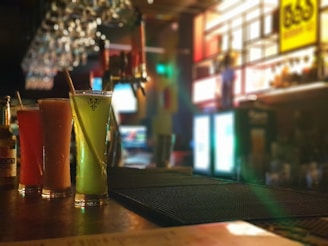 Three tall glasses filled with colorful beverages are arranged on a bar counter. The background features dim ambient lighting, with glowing signs and a row of hanging glasses creating a cozy atmosphere. The focus on the drinks contrasts with the blurred background.