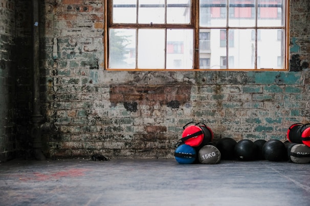 A rustic gym setting with an exposed brick wall and a large window. Several medicine balls, some marked with weights like 15kg, are stacked against the wall. The space conveys an industrial and minimalistic vibe.