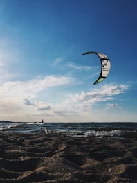 A kite surfer glides over ocean waves beneath a clear blue sky, with a sandy beach in the foreground. The kite is prominently visible in the sky, while the surfer is silhouetted against the horizon.