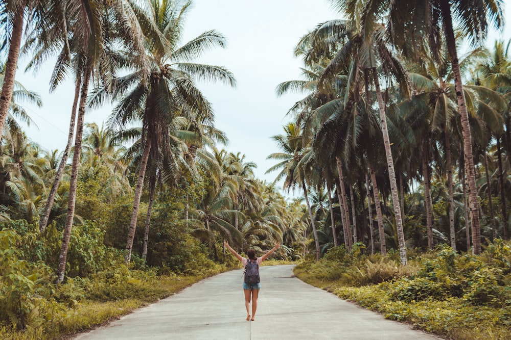 woman walking in the middle of the road raising both her arms surrounded by grass and coconut trees