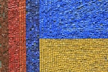 A vibrant mosaic composed of small, square tiles arranged in geometric patterns. The mosaic features three main sections: a vertical strip of red tiles, a horizontal section of blue tiles, and a block of yellow tiles. Each section consists of tiles slightly varying in shade, creating a textured and dynamic effect.