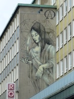 A mural on the side of a building depicting a serene, ethereal woman holding a candle. She has long hair and closed eyes, wearing flowing robes. Intricate designs and symbols surround her, including an Alpha and Omega sign above her head. The artwork blends seamlessly into the architecture with muted tones.