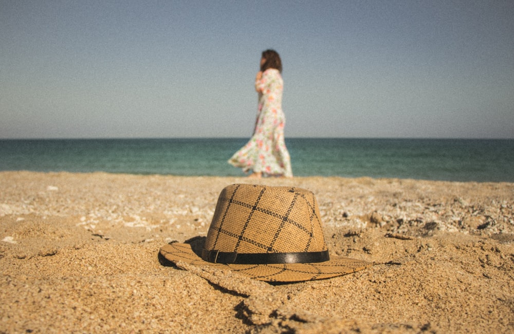 mujer caminando cerca de la orilla del mar y sombrero marrón y gris sobre arena marrón