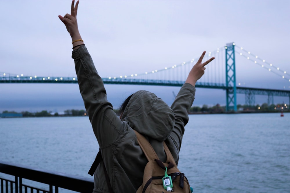 person wears hoodie and backpack near body of water