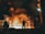 Workers stand near a large industrial furnace with bright flames and heavy smoke, suggesting a metal or steel manufacturing process. The lighting is dim with an orange glow illuminating the scene, enhancing the industrial atmosphere.