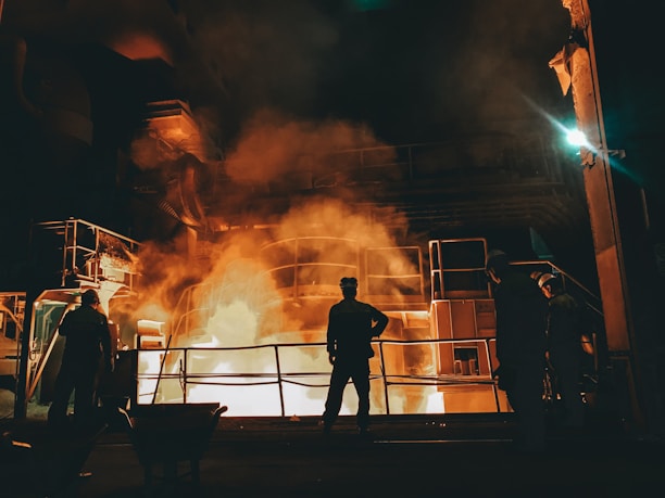 Workers stand near a large industrial furnace with bright flames and heavy smoke, suggesting a metal or steel manufacturing process. The lighting is dim with an orange glow illuminating the scene, enhancing the industrial atmosphere.