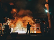 Workers stand near a large industrial furnace with bright flames and heavy smoke, suggesting a metal or steel manufacturing process. The lighting is dim with an orange glow illuminating the scene, enhancing the industrial atmosphere.