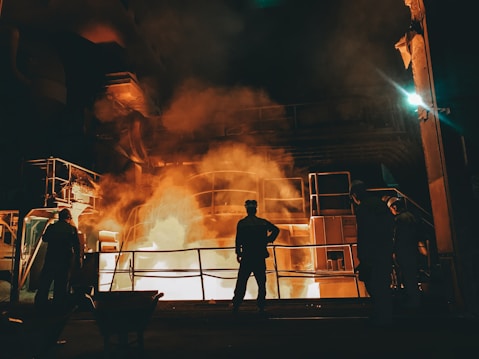Workers stand near a large industrial furnace with bright flames and heavy smoke, suggesting a metal or steel manufacturing process. The lighting is dim with an orange glow illuminating the scene, enhancing the industrial atmosphere.