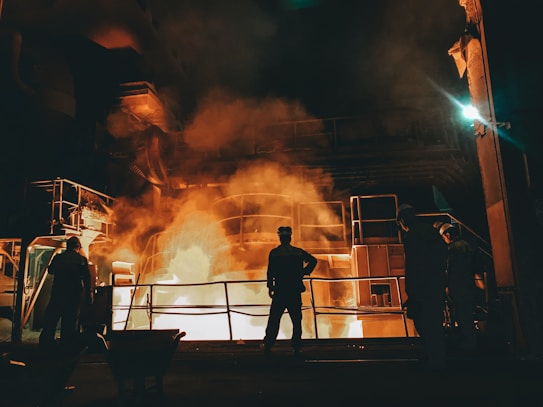 Workers stand near a large industrial furnace with bright flames and heavy smoke, suggesting a metal or steel manufacturing process. The lighting is dim with an orange glow illuminating the scene, enhancing the industrial atmosphere.