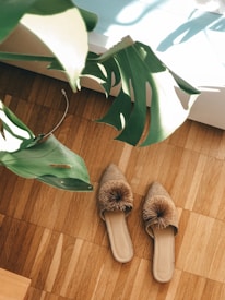 A pair of straw-like slippers with decorative pom-poms lies on a light wooden floor. Large green monstera leaves are partially visible in the top left corner, with sunlight streaming through and casting shadows.