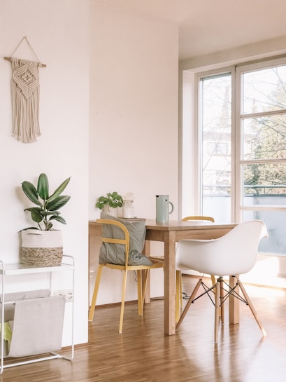 A bright and minimalist interior space featuring a wooden dining table with two chairs, one white and one yellow. A potted plant sits on the table, along with a jug and a light bulb decoration. The room is illuminated by natural light from a large window. A macram&eacute; wall hanging decorates the white wall, and a small rack with a basket is placed nearby.