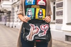A person is holding a black, graphic handbag featuring artistic depictions of high-heeled legs, a lipstick, and a heart. The individual is wearing a colorful t-shirt with illustrations of women and slogans. The background consists of a city street with architectural details of buildings.