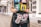 A person is holding a black, graphic handbag featuring artistic depictions of high-heeled legs, a lipstick, and a heart. The individual is wearing a colorful t-shirt with illustrations of women and slogans. The background consists of a city street with architectural details of buildings.