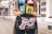 A person is holding a black, graphic handbag featuring artistic depictions of high-heeled legs, a lipstick, and a heart. The individual is wearing a colorful t-shirt with illustrations of women and slogans. The background consists of a city street with architectural details of buildings.