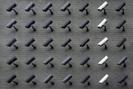 A grid pattern of surveillance cameras is mounted on a gray brick wall. Most of the cameras are black, with a few white ones interspersed, all angled uniformly to the left.