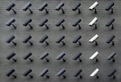 A grid pattern of surveillance cameras is mounted on a gray brick wall. Most of the cameras are black, with a few white ones interspersed, all angled uniformly to the left.