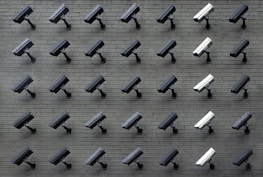 A grid pattern of surveillance cameras is mounted on a gray brick wall. Most of the cameras are black, with a few white ones interspersed, all angled uniformly to the left.
