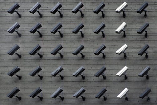 A grid pattern of surveillance cameras is mounted on a gray brick wall. Most of the cameras are black, with a few white ones interspersed, all angled uniformly to the left.