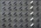 A grid pattern of surveillance cameras is mounted on a gray brick wall. Most of the cameras are black, with a few white ones interspersed, all angled uniformly to the left.