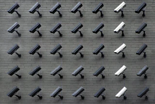A grey brick wall covered in a dense grid of black security cameras, with one vertical column of pale cameras standing out
