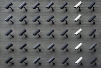 A grid pattern of surveillance cameras is mounted on a gray brick wall. Most of the cameras are black, with a few white ones interspersed, all angled uniformly to the left.