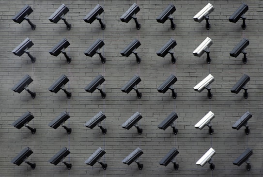 A grid pattern of surveillance cameras is mounted on a gray brick wall. Most of the cameras are black, with a few white ones interspersed, all angled uniformly to the left.