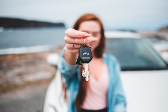 A person is holding car keys in focus while standing in front of a blurred car and a scenic background of water and hills. The keys have a distinctive logo on the keychain. The person's face and upper body are out of focus, highlighting the keys.