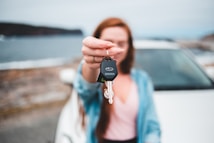 A person is holding car keys in focus while standing in front of a blurred car and a scenic background of water and hills. The keys have a distinctive logo on the keychain. The person's face and upper body are out of focus, highlighting the keys.