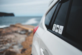 A white car is parked near a coastal area with the ocean and a rocky shoreline in the background. The car's side mirrors and window bear several stickers featuring minimalist icons.