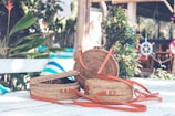 A pair of woven wicker handbags with leather straps are placed on a white outdoor table. Surrounding the table are various lush green plants, with a blue and white nautical theme visible in the background, including a decorative ship's wheel.