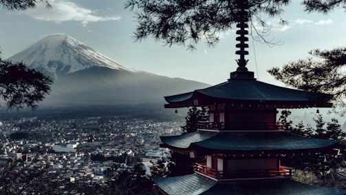 A traditional Japanese pagoda stands prominently in the foreground, nestled among pine trees. In the background, Mount Fuji rises majestically under a sky with light clouds, overlooking a sprawling city below.