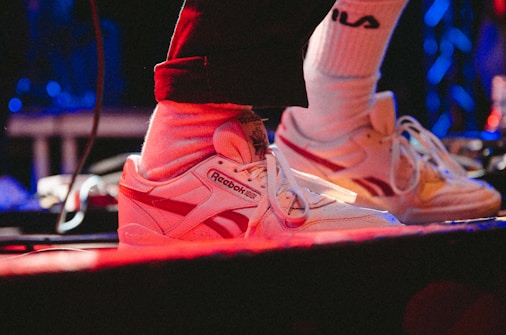 A pair of white Reebok sneakers with red accents is worn with white socks and dark pants. Bright red and blue stage lights create a vibrant atmosphere, with cables visible around the feet.