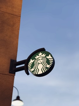 A circular outdoor sign features the iconic green logo against the backdrop of a clear sky. The sign is mounted on a rust-colored wall, with part of a street lamp visible below.