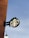 A circular outdoor sign features the iconic green logo against the backdrop of a clear sky. The sign is mounted on a rust-colored wall, with part of a street lamp visible below.