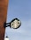 A circular outdoor sign features the iconic green logo against the backdrop of a clear sky. The sign is mounted on a rust-colored wall, with part of a street lamp visible below.