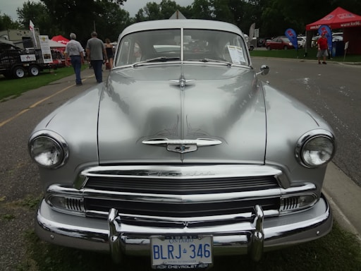 A classic silver Chevrolet car with a vintage design is parked on a street at an outdoor event. The front grille and headlights are prominent, and the car's license plate is visible. Several people are nearby, and there are red and blue tents in the background with some cars parked further away.