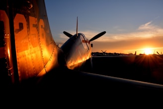 A vintage airplane is parked on a runway during sunset, with the light casting a warm glow on its metallic surface. The silhouette of its propeller and tail are prominent against the sky, while other aircraft are faintly visible in the background.