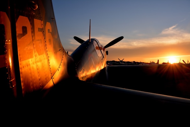 A vintage airplane is parked on a runway during sunset, with the light casting a warm glow on its metallic surface. The silhouette of its propeller and tail are prominent against the sky, while other aircraft are faintly visible in the background.