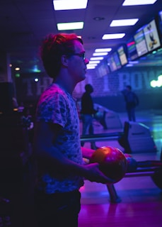 A person is holding a bowling ball, standing in a neon-lit bowling alley. The environment is dimly lit with an array of colorful lights reflecting off surfaces, creating a vibrant atmosphere. Multiple screens are visible in the background, displaying information. There are other people in the alley, some of whom are engaged with the bowling lanes.